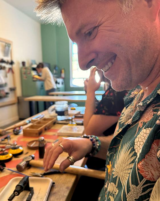 A person smiling at a workbench crowded with jewelry-making tools