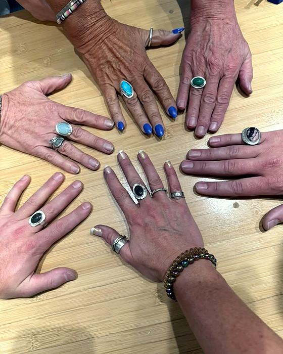 A circle of hands resting together on a wooden table, each wearing a silver ring set with a stone