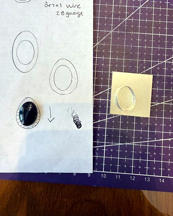 Hand-drawn bezel sketches with notes beside a cabochon and a small sheet of silver on a cutting mat