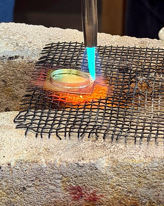 A torch flame heating a silver bezel on a soldering block covered with wire mesh