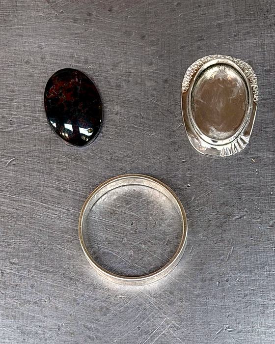 A cabochon, a finished bezel cup, and a plain silver band laid out on a steel block